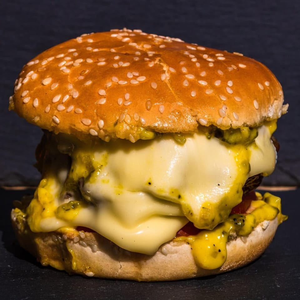 A Samz Smash burger with melted cheese and a yellow sauce oozing out, on a dark surface.